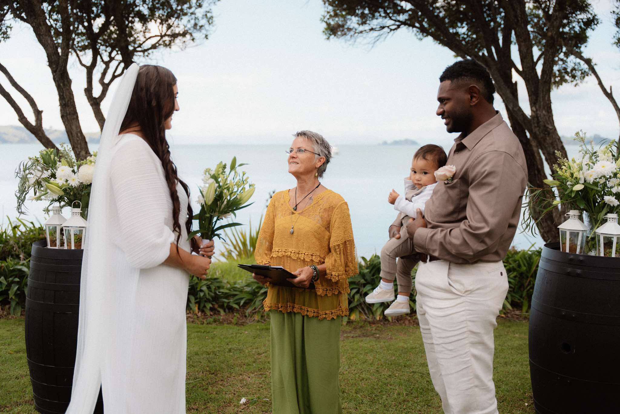 Relaxed garden wedding ceremony on Waiheke Island - couple with baby and celebrant, harbour in background